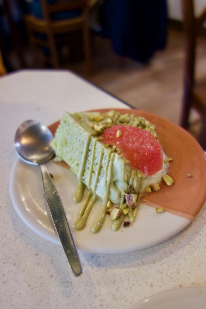 Part de bûche pistache avec des pistaches et une tranche de pamplemousse sur dessus, accompagnée d'une cuillère, le tout dans une assiette blanche et ocre