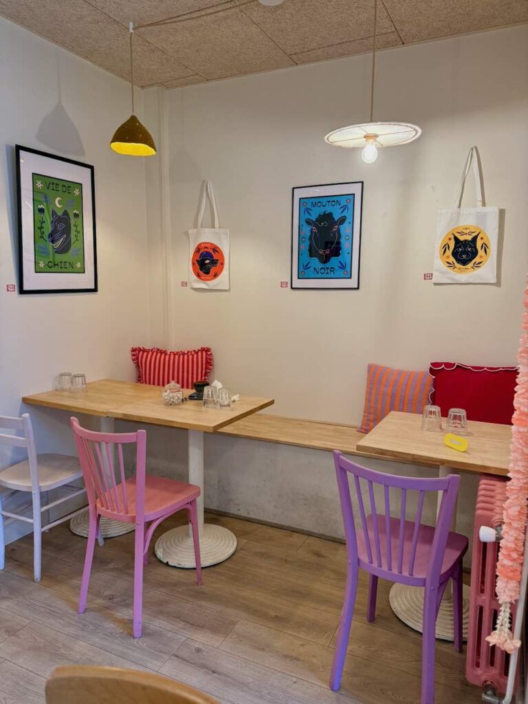 Intérieur de Fricotte avec deux tables en bois, des chaises et coussins roses et rouges, tableaux d'artiste au mur