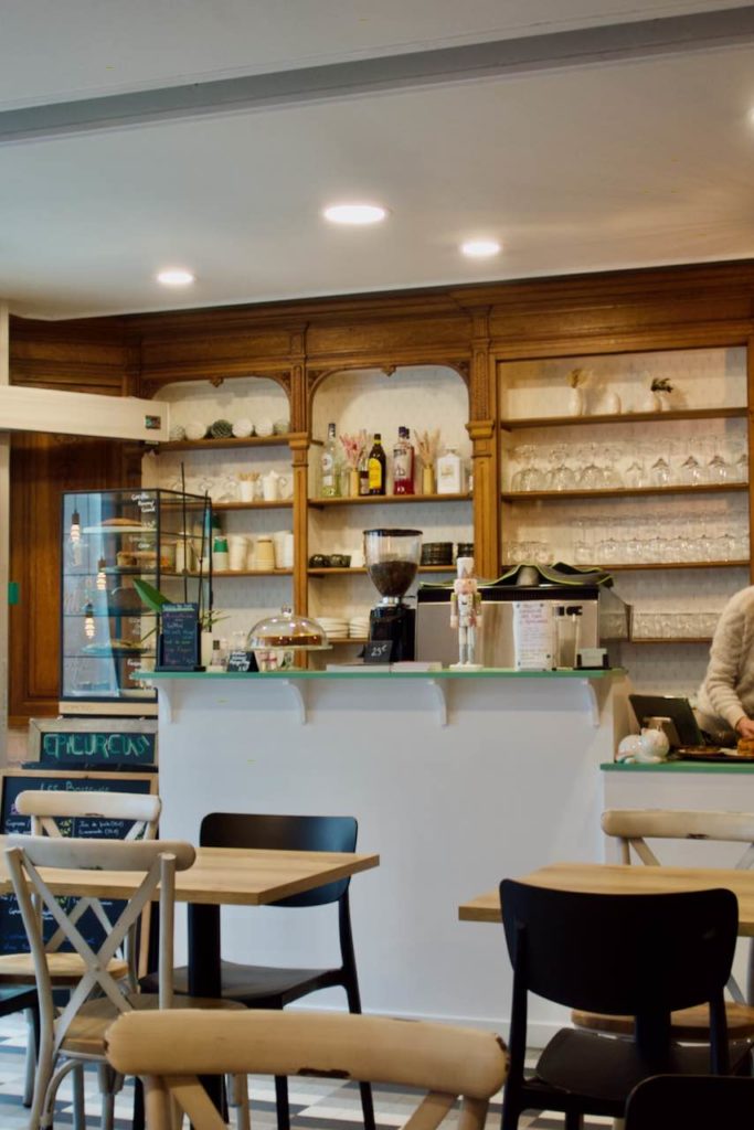 Intérieur du café avec au premier plan des chaises et tables en bois, au second plan le comptoir blanc et au dernier plan des étagères en bois au mur avec des verres et des bouteilles