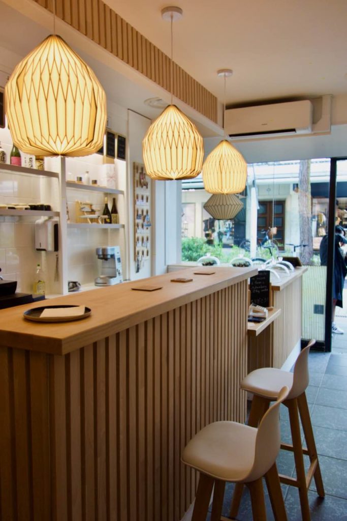 Bar en bois façon claustras, luminaires en bois clair, murs blancs avec de petites étagères sur lesquelles sont posés des sakés