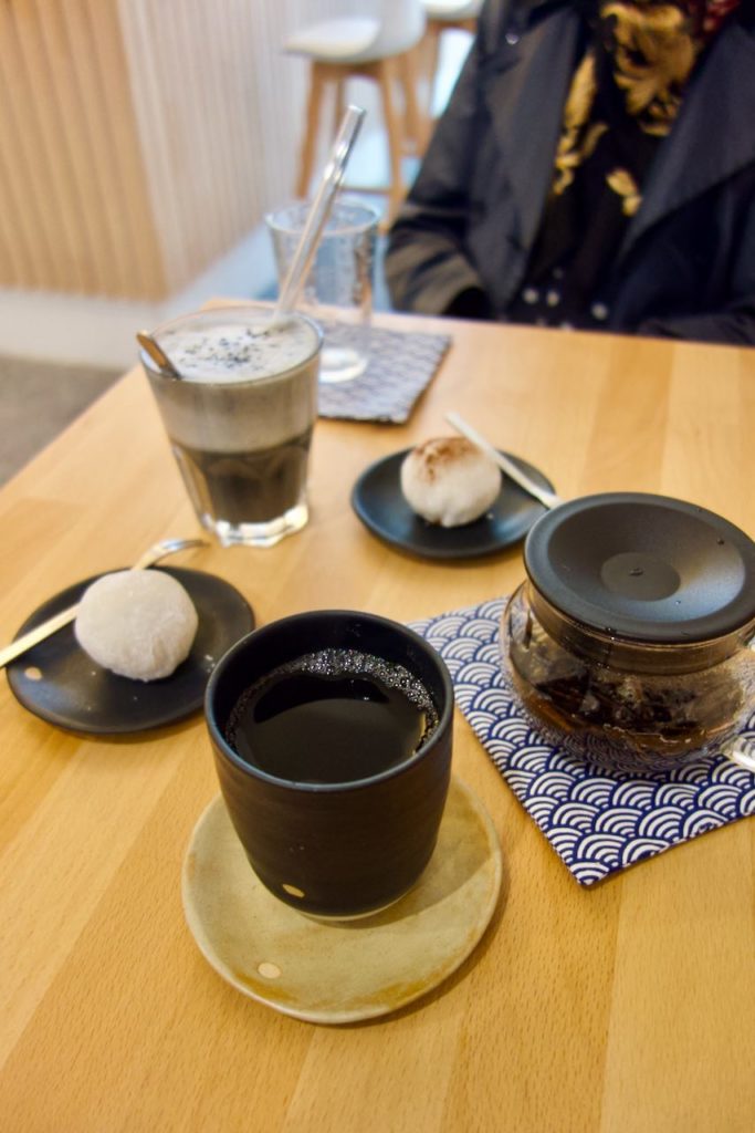 Tasse de thé, théière individuelle, deux mochis sur des coupelles noires et verre de black sésame latté sur une table en bois. Accompagnés de deux serviettes à pattern vagues japonaises.