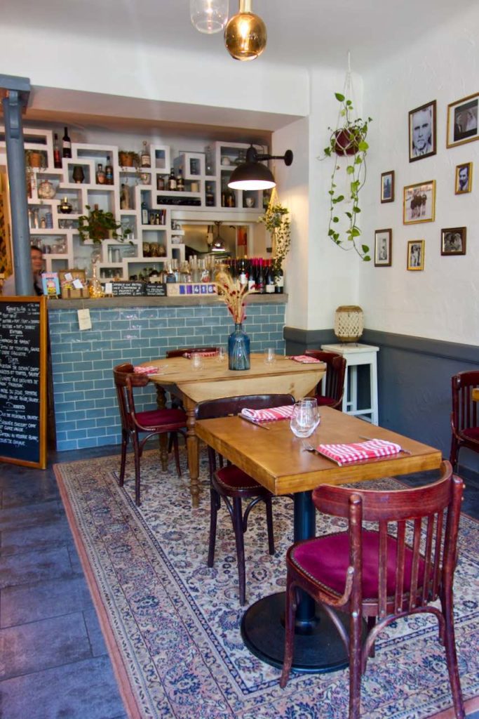 Intérieur du restaurant avec une table ronde en bois à bords rabattus, un comptoir avec crédence bleue, des cadres au mur, un large tapis au sol