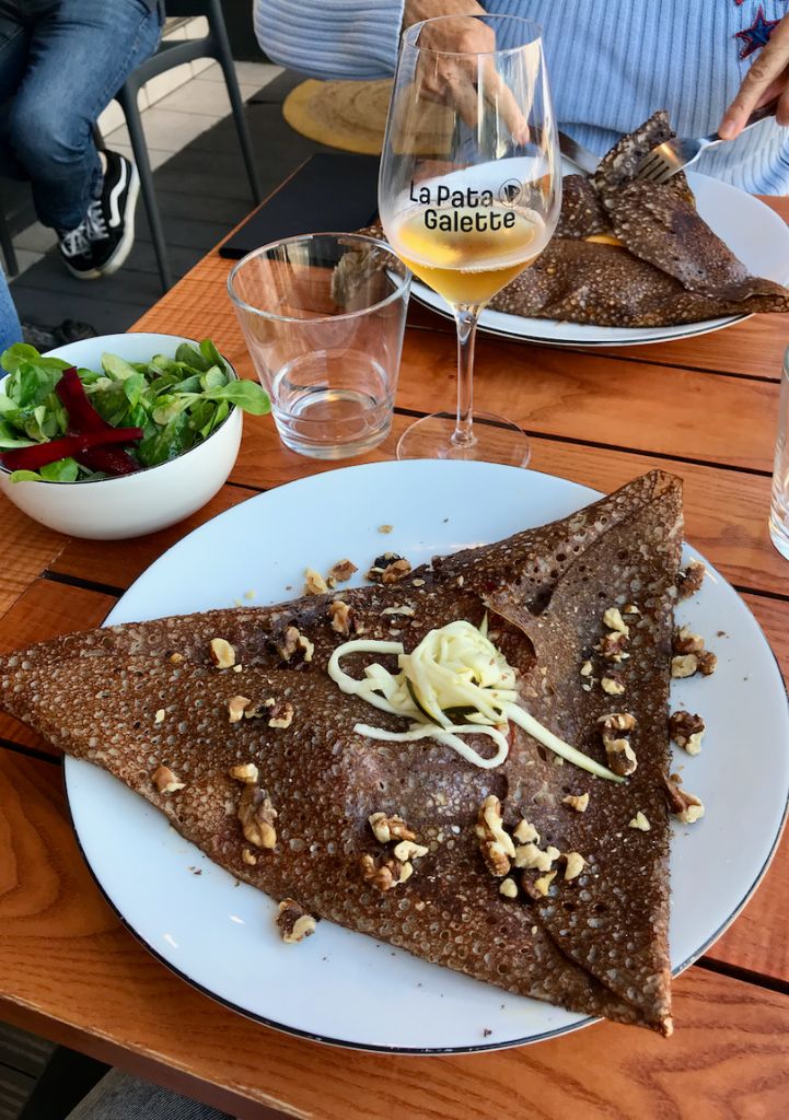 Deux galettes dans des assiettes blanches, un verre de cidre, un bol de salade sur une table en bois