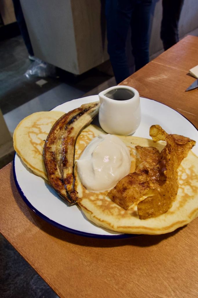Assiette de pancakes, banane flambée, bacon végétal, pot de sirop d'érable dans une assiette blanche