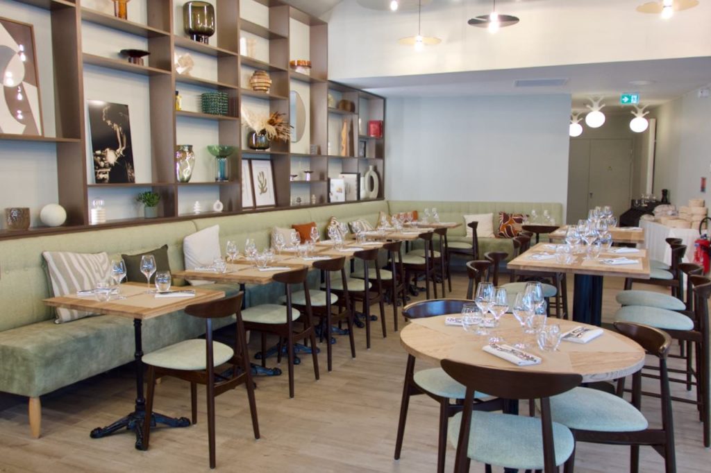 Intérieur de l'Atelier avec une longue banquette anis accueillant plusieurs tables en bois. Des étagères en bois au mure, parquet et chaises en bois noires.