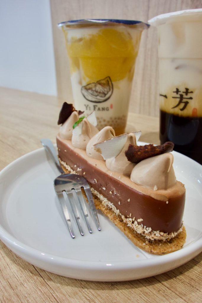 Gâteau individuel dans une assiette blanche avec une fourchette et de bubble teas en arrière plan