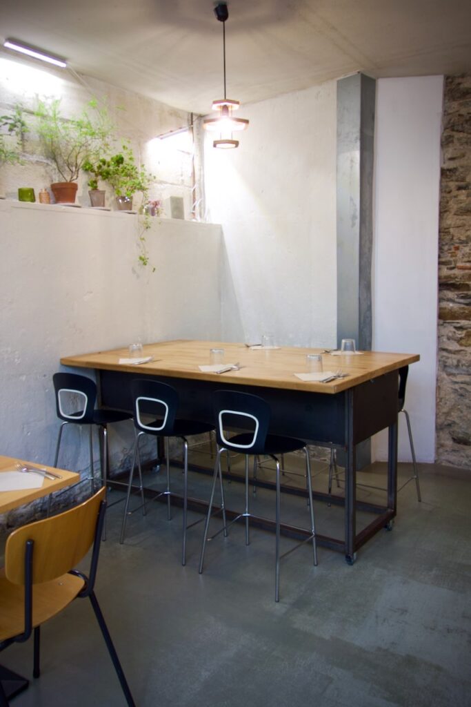Table haute en bois, chaises hautes noires, mur blanc sur lequel sont posés des pots de plantes aromatiques