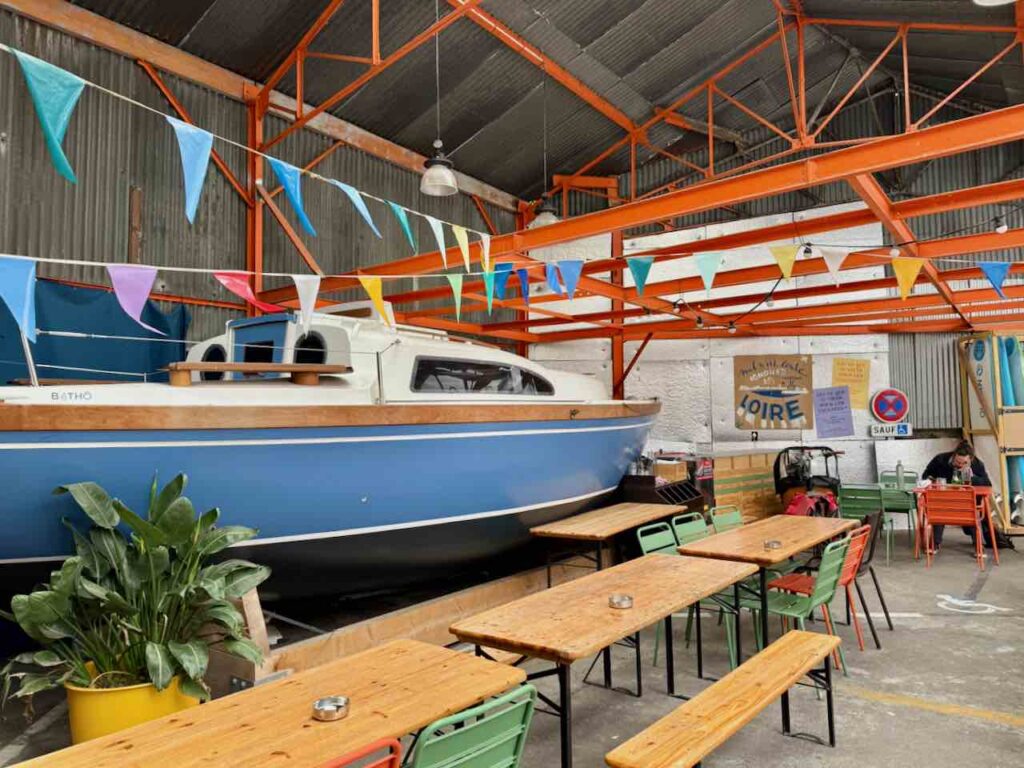 Cour intérieure du Lieu-Dit avec de longues tables et bancs en bois ainsi qu'un bateau bleu et blanc en arrière plan