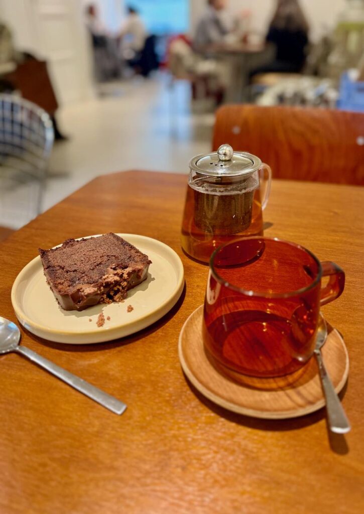 Gâteau au chocolat dans une assiette blanche à gauche, tasse vide ocre sur une coupelle en bois à droite, petite théière en verre individuelle au second plan