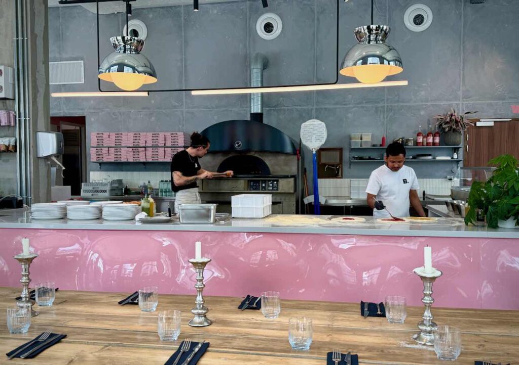 Vaste comptoir rose brillant derrière lequel deux personnes préparent des pizza.