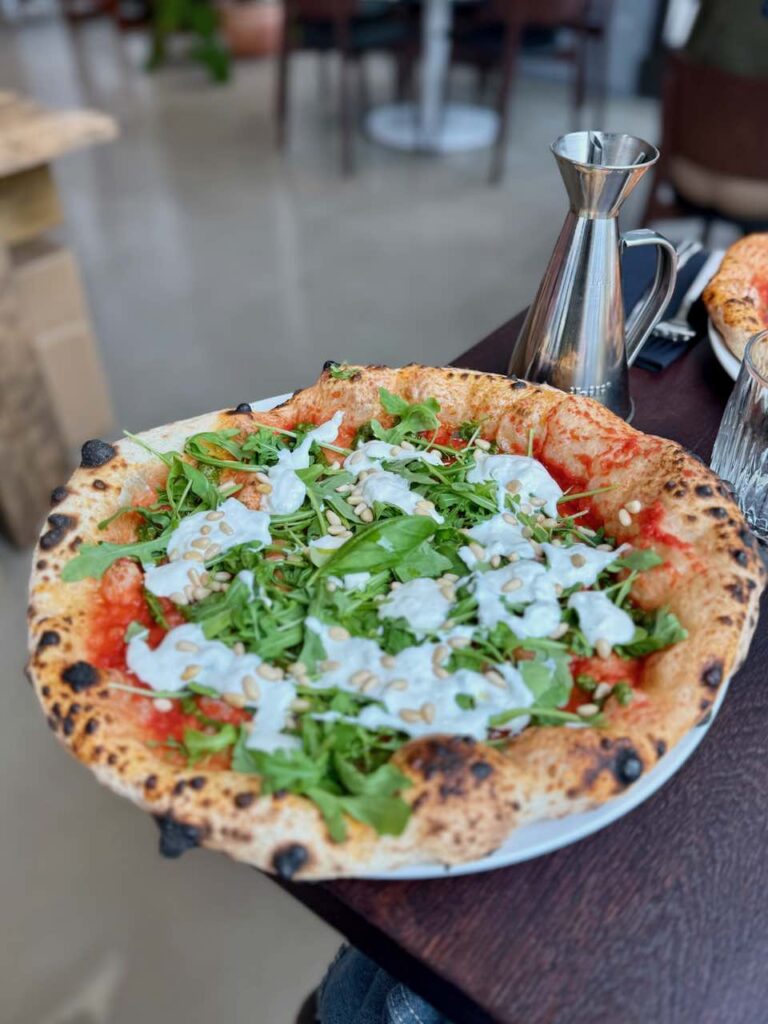 Pizza parsemée de stracciatella et de roquette, un contenant en métal de sauce piquante au second plan