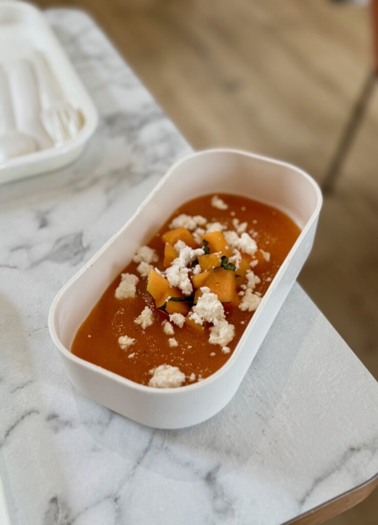 Soupe de melon parsemée de chèvre frais dans un bento blanc rectangulaire sur une table effet marbre