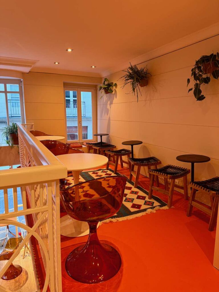 Vue de l'espace café au 1er étage avec chaises design plastique orange transparent, tables rondes blanches et petites tables accrochées au mur, grand tapis au sol et sol orange