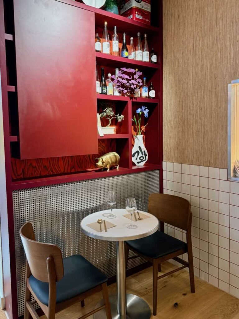 Détail d'une table bistro avec deux chaises design en bois, carreaux de faïence blancs au mur, étagère rouge d'inspiration coréenne