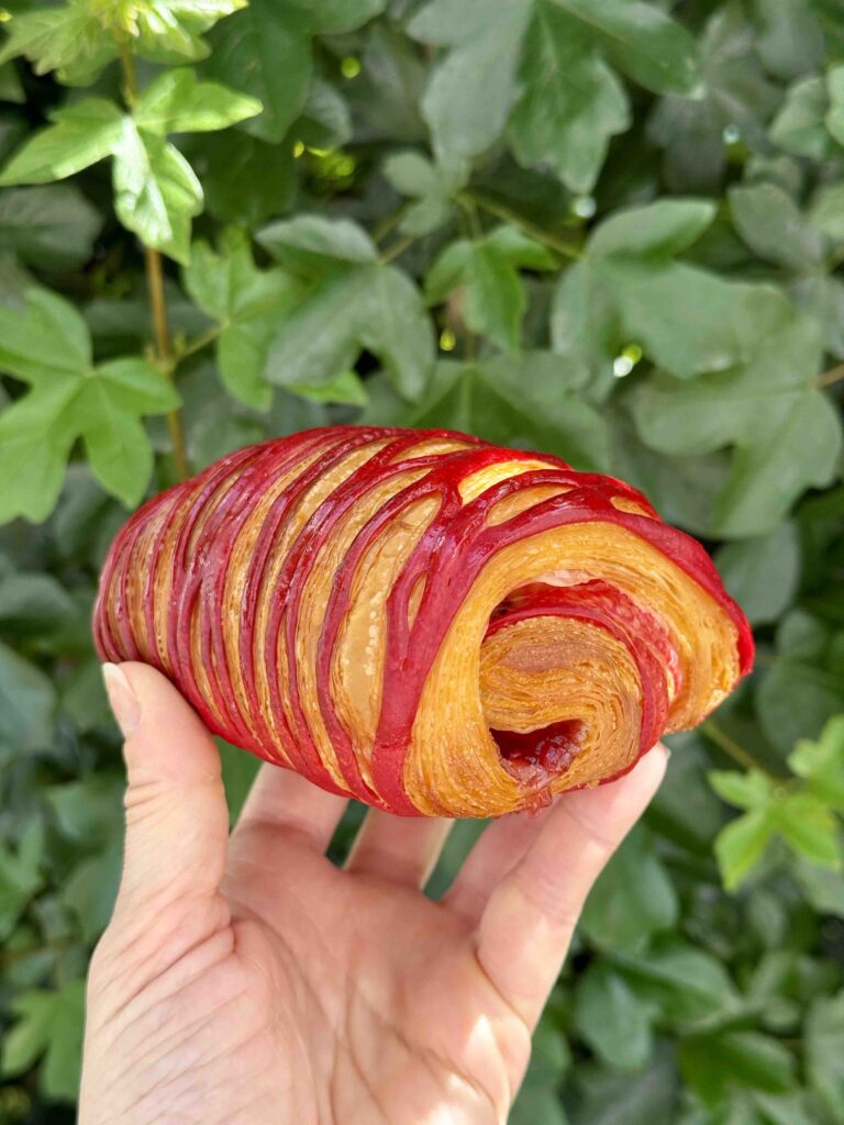 Pain au chocolat avec des stries graphiques roses tenu dans la main devant un arbre