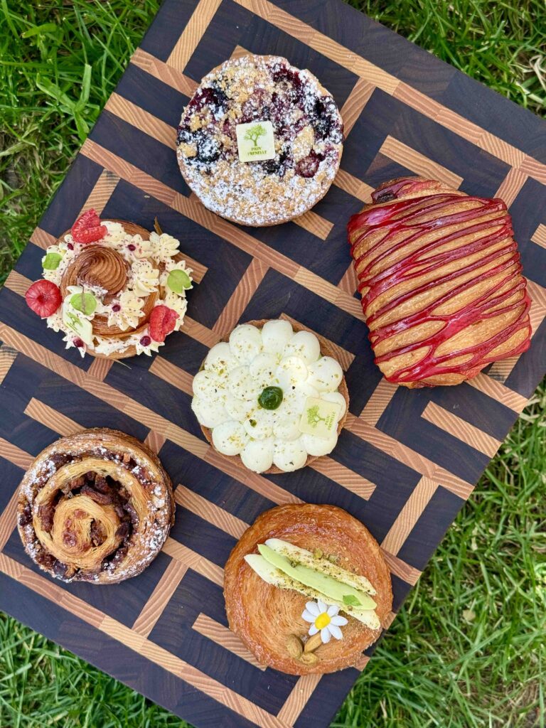 Six patisseries et viennoiseries très colorées photographiées du dessus sur une planche en bois aux lignes graphiques et posée dans l'herbe