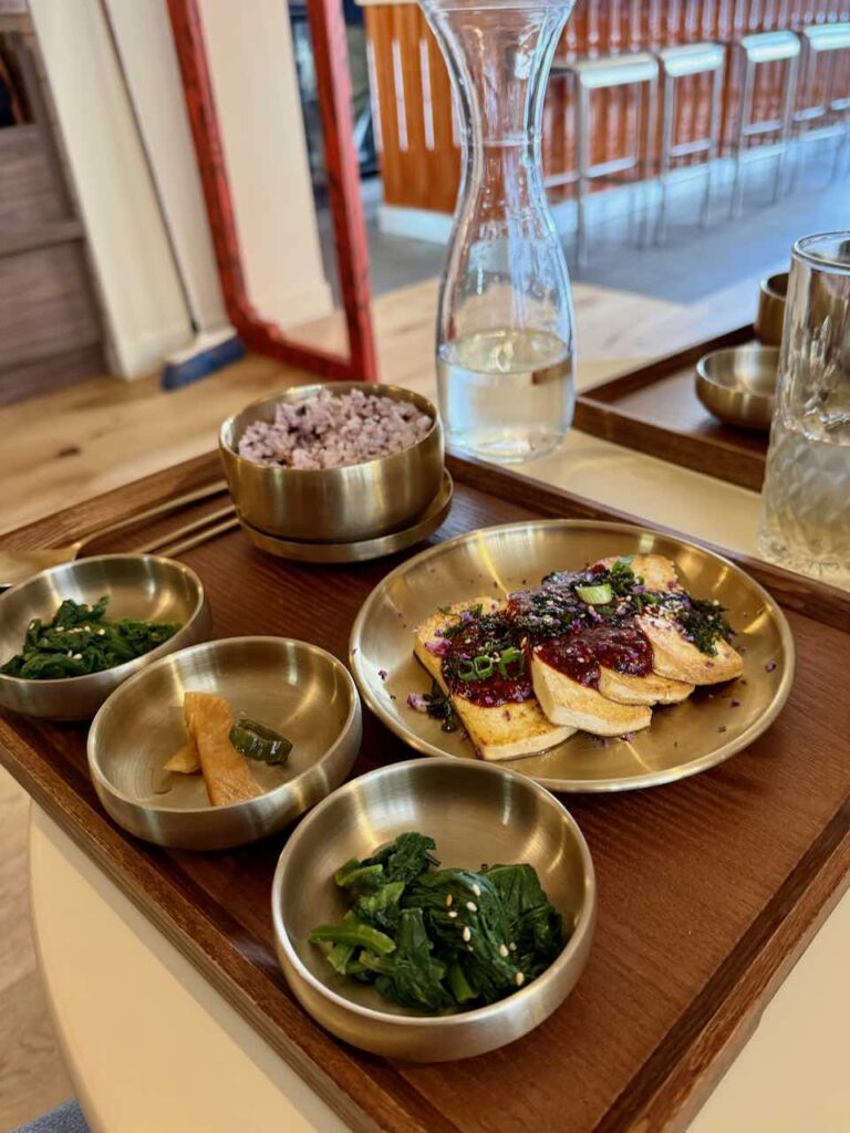 Tofu frit dans une assiette dorée avec un trio d'accompagnements dans de petits bols ronds en métal, bol de riz rouge sur un plateau en bois sombre