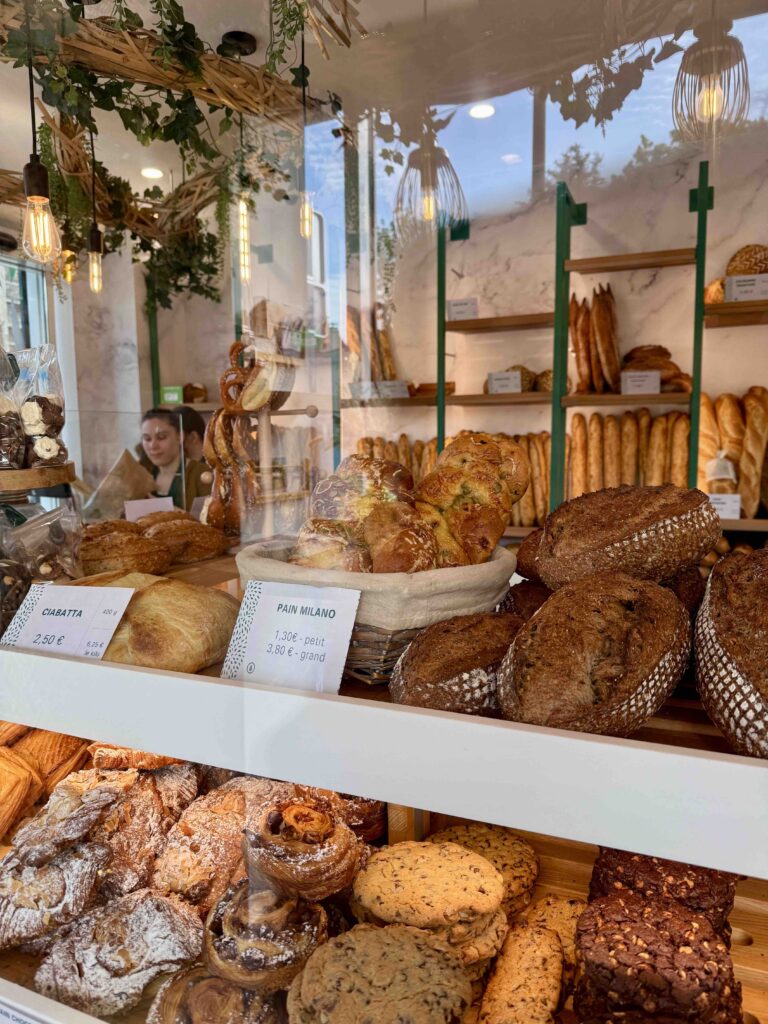 Pains et viennoiseries sur deux niveaux dans la vitrine, noms et pains affichés, baguette sur les étagères au second plan