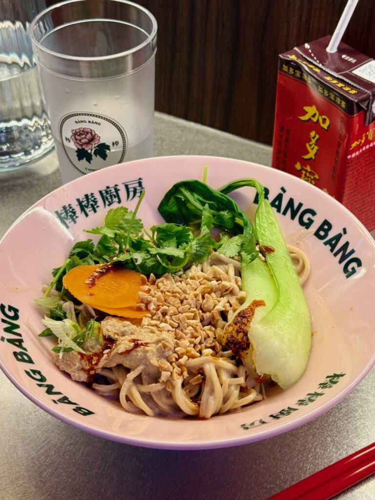 Nouilles, pak choi entier, tranches de carotte, coriandre fraîche et cacahuètes dans un bol blanc à l'effigie du restaurant