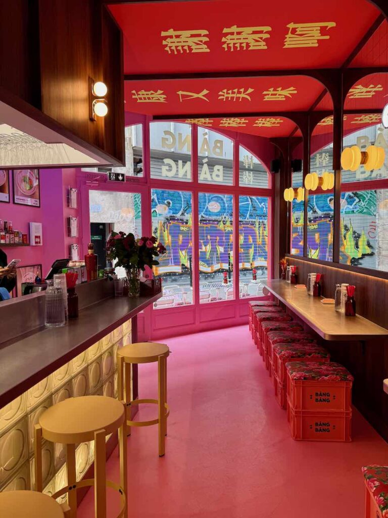 Intérieur tout en longueur de Bang Bang, zinc et tabourets hauts à gauche, comptoir et tabourets à base de caisses en plastique sur lesquels sont posés des poufs à droite, entrée dans le fond avec vue sur un tag
