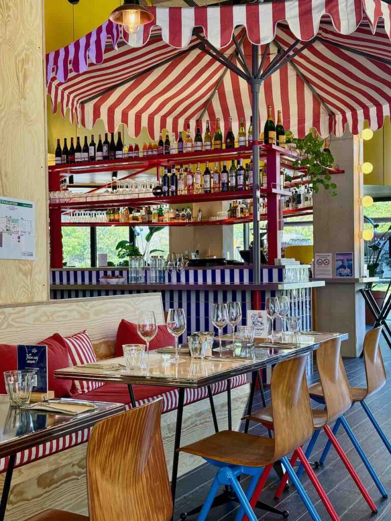 Tables bistro, chaises en bois, banquettes bois et coussin rouge à rayures blanches, zinc bleu à rayures blanches et bouteilles sur des étagères rouges