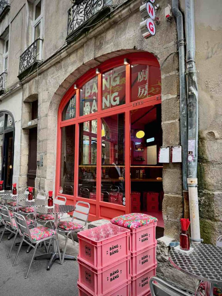Devanture de Bang Bang : une large porte en arcade aux huisseries rouge, terrasse avec chaises et tables bistro devant