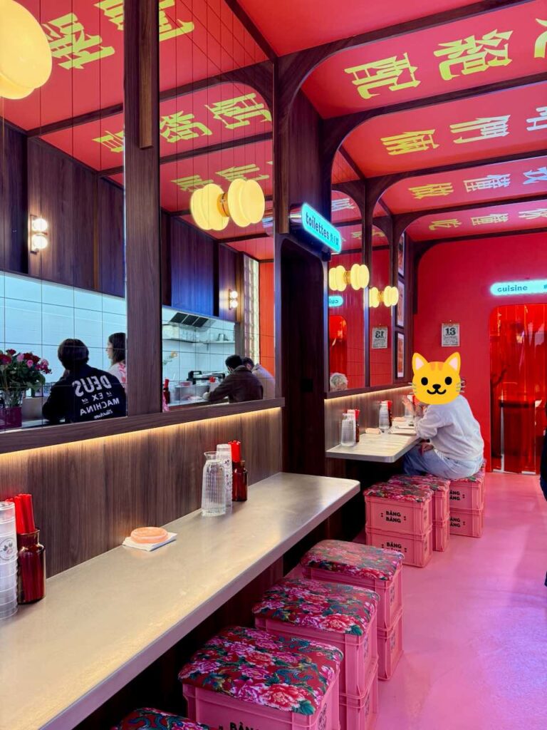 Vue opposée, toujours en longueur avec gros plan sur le comptoir en métal gris et les tabourets caisse en plastique rose