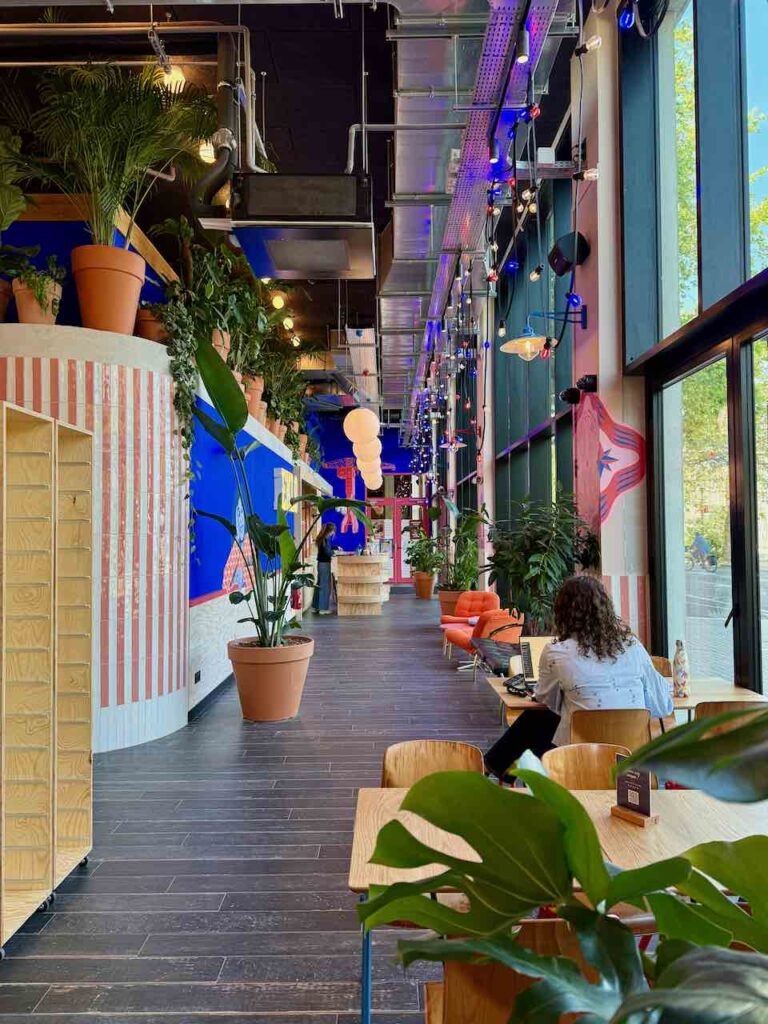 Couloir aux murs colorés avec quelques tables en bois, plusieurs plantes en pot et des fauteuil corail