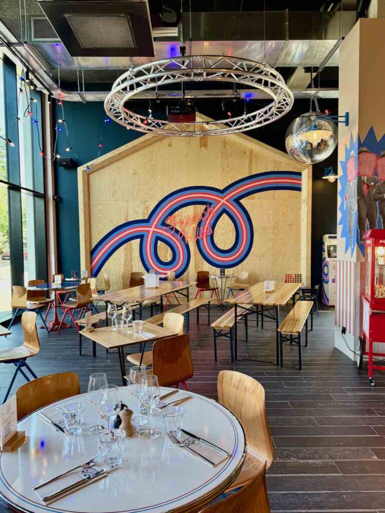 Table bistro et verre au premier plan, tables et longs bancs en bois au second plan, logo de French Kiss au mur au dernier plan