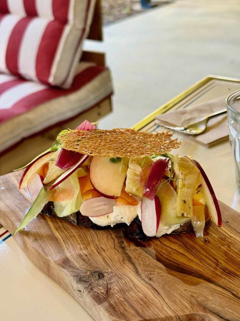 Tartine généreusement surmontée de légumes et fruits sur lesquels est déposée une tuile de parmesan, le tout sur une planche en bois