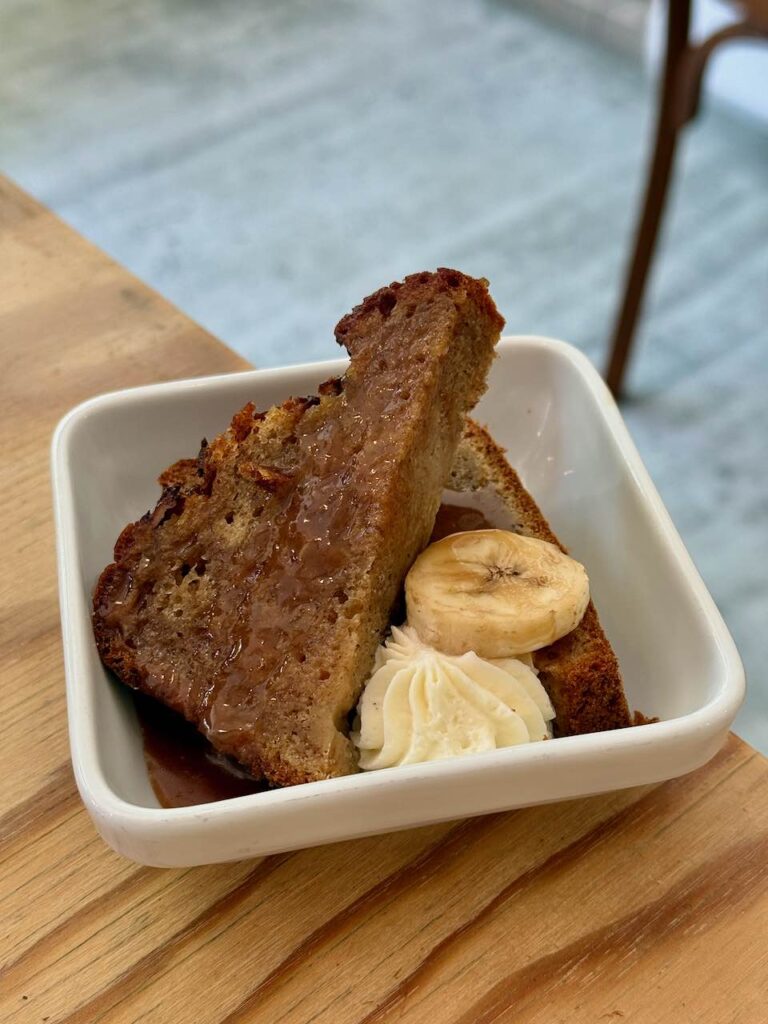 Tranche de banana bread coupée en deux, arrosée de caramel au beurre salé, avec une tranche de banane et une pointe de chantilly dans un récipient carré blanc à bords hauts
