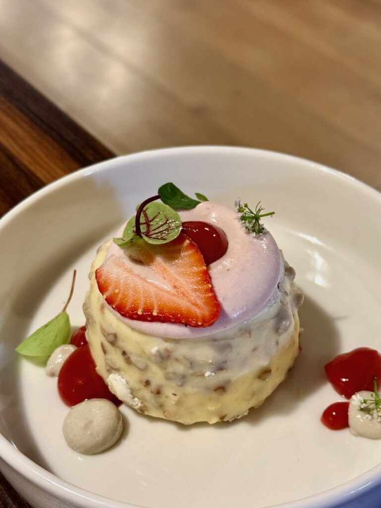 Nougat chocolat blanc surmonté d'une crème fraise, d'une tranche de fraise, avec pointes de gel autour dans une assiette blanche à bords hauts
