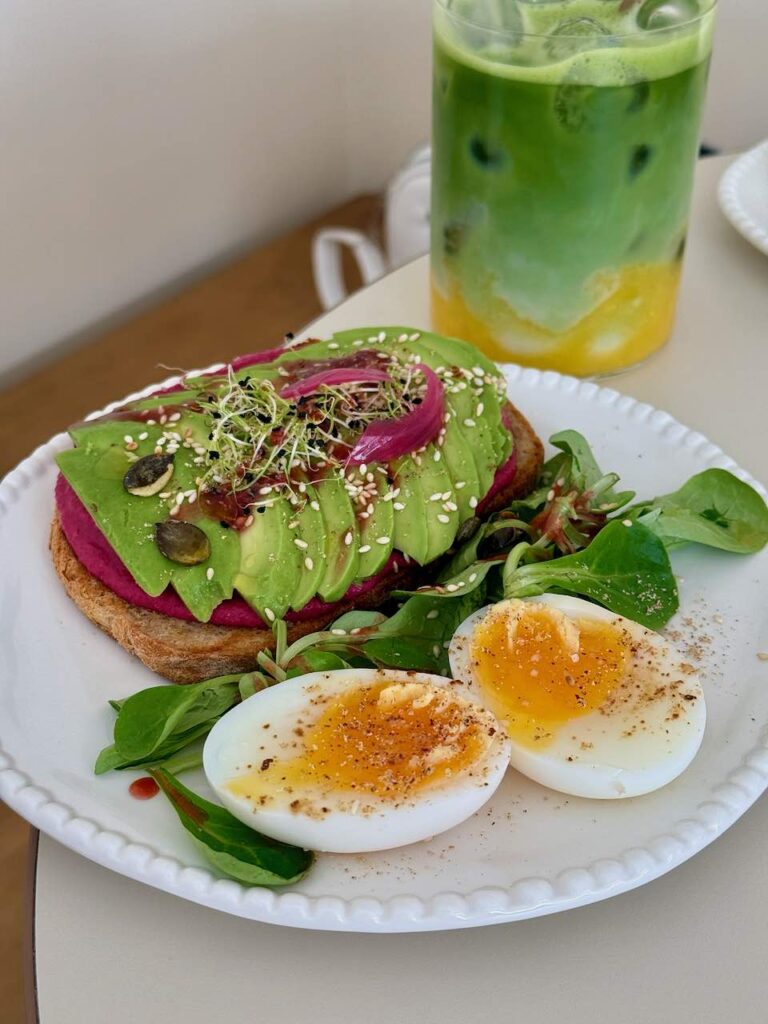 Tranches d'avocat sur du houmous rose, lui-même sur une tranche de pain, oeuf mollet coupé en deux, le tout dans une assiette blanche