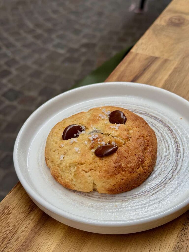 Cookie avec trois palets de chocolat au lait et surmonté de fleur de sel dans une assiette en grès plate à bords hauts