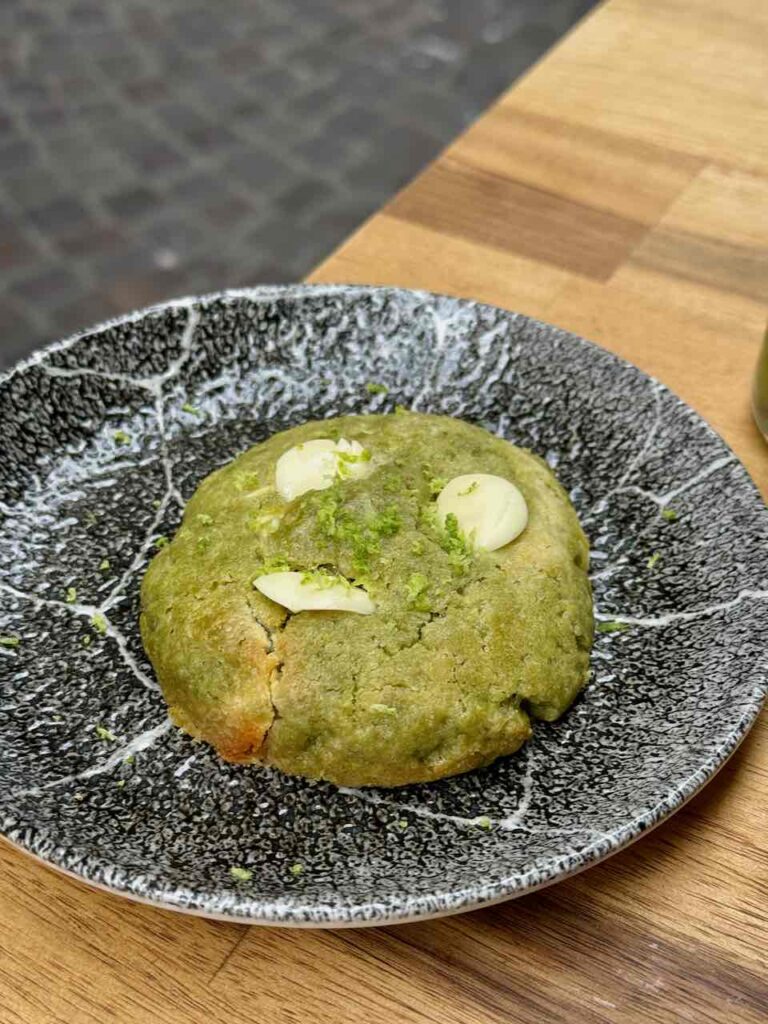 Cookie matcha avec trois palets de chocolat blanc, surmonté de zestes de citron vert dans une assiette noire et blanche