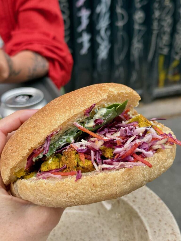 Sandwich tenu à la main laissant apparaître des falafels, du chou rouge, des carottes et de la salade